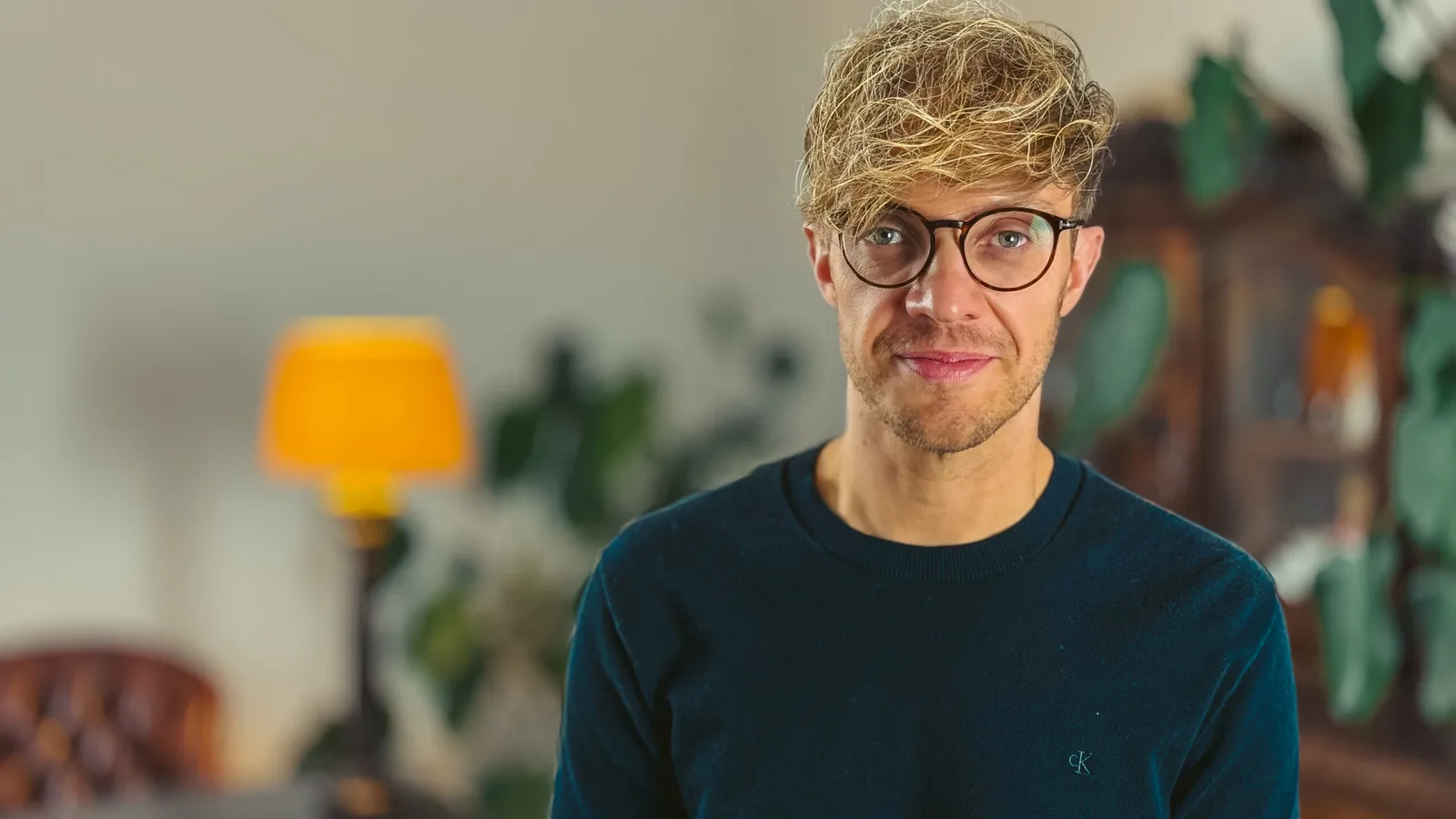 Mann mit blondem Haar und Brille in dunklem Hemd vor unscharfem Hintergrund mit Lampe
