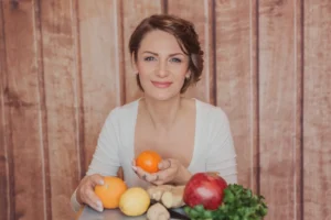 Frau mit braunem Haar in weißem Shirt hält Orangen und Apfel, gesunde Lebensmittel auf Tisch, Body Balance Hormonbasiertes Ab