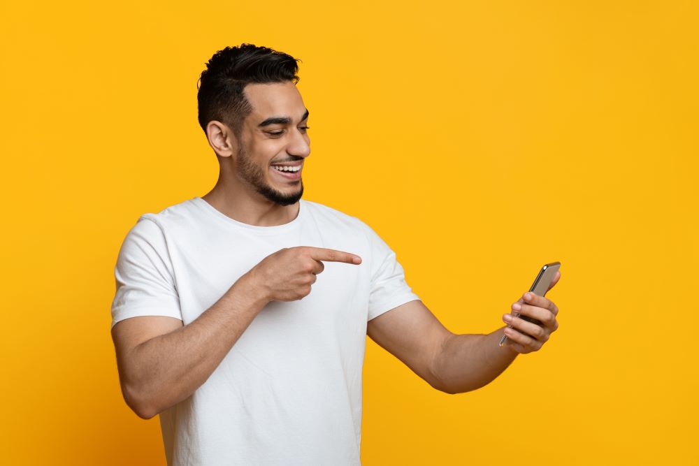 Mann im weißen T-Shirt auf gelbem Hintergrund hält Smartphone, deutet darauf hin