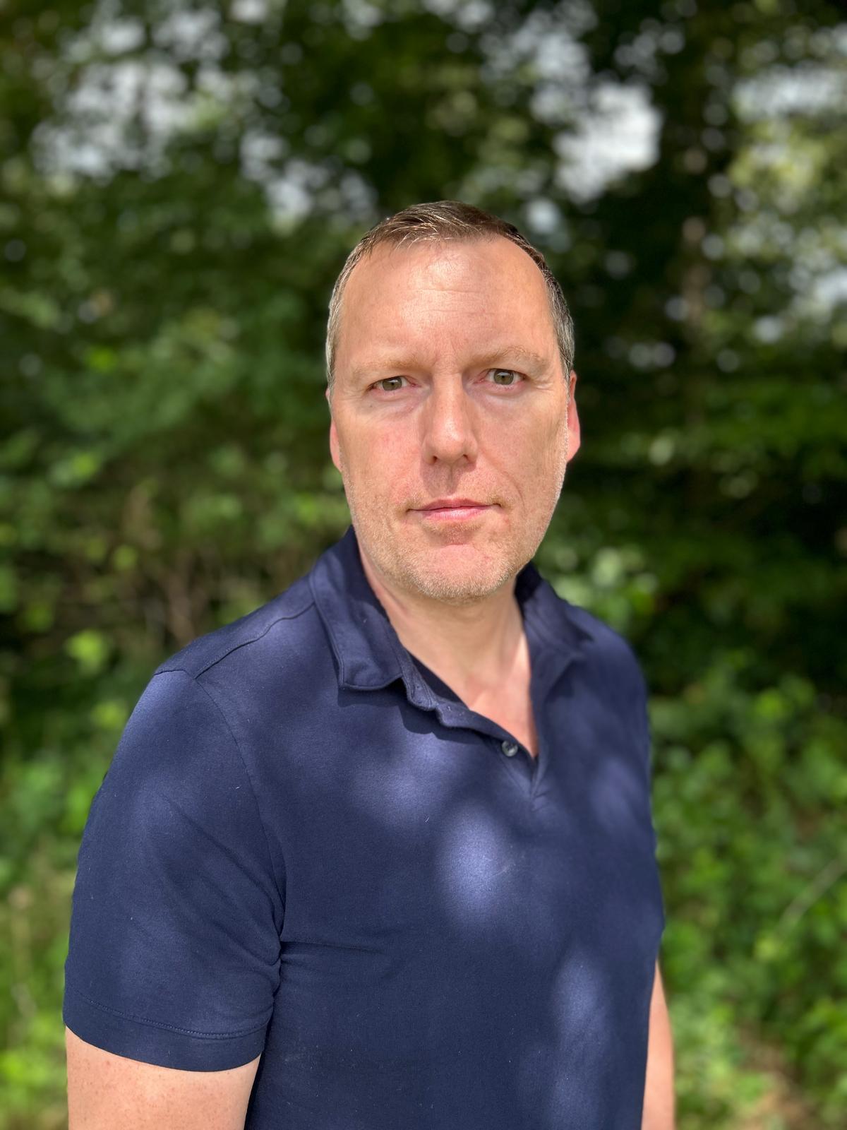 Porträtfoto eines Mannes mittleren Alters in dunklem Poloshirt vor Grünflächenhintergrund