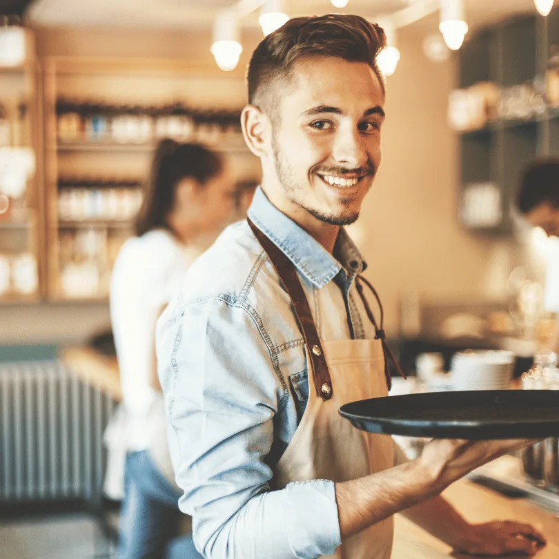 Junger Kellner in hellem Hemd mit Tablett im modernen Bistro mit Gästen im Hintergrund