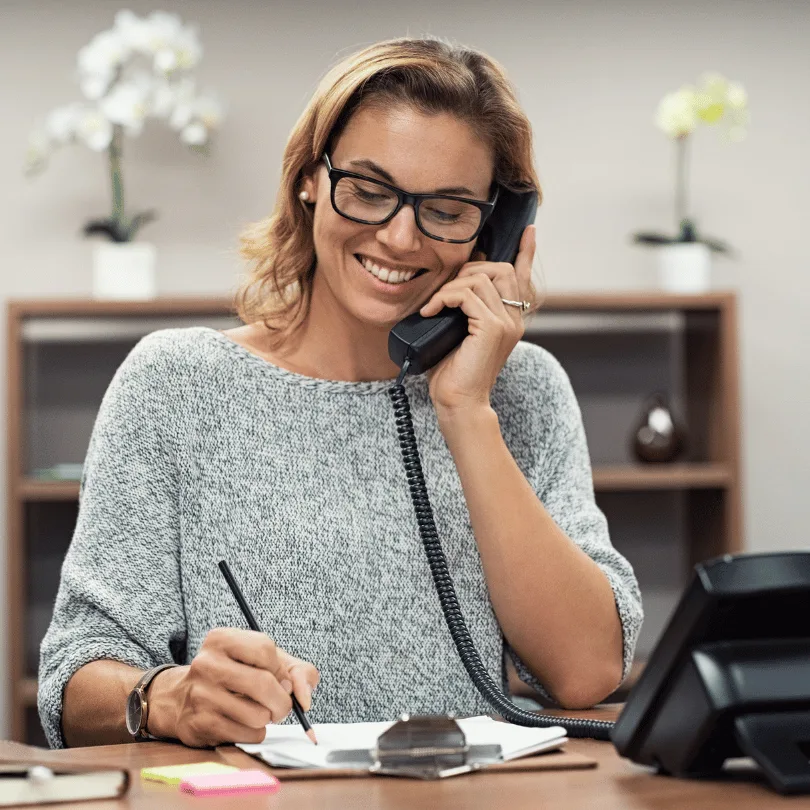 Frau mit Brille am Schreibtisch telefonierend, professionelle Büroumgebung mit Blumen und Regalen