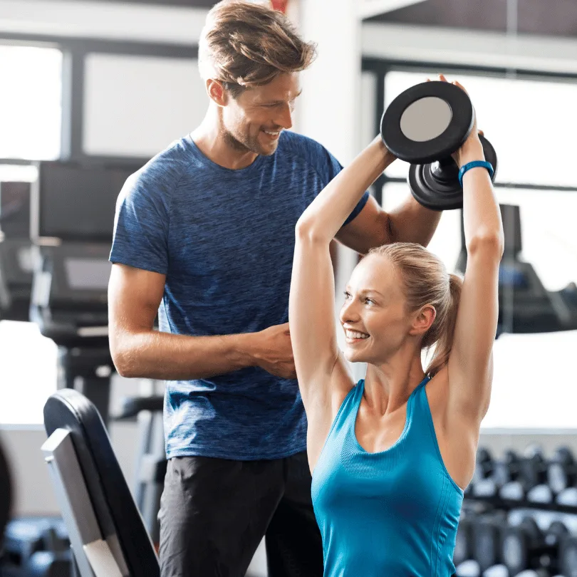 Personal Trainer mit Kunde beim Hantelstemmen in modernem Fitnessstudio mit großen Fenstern