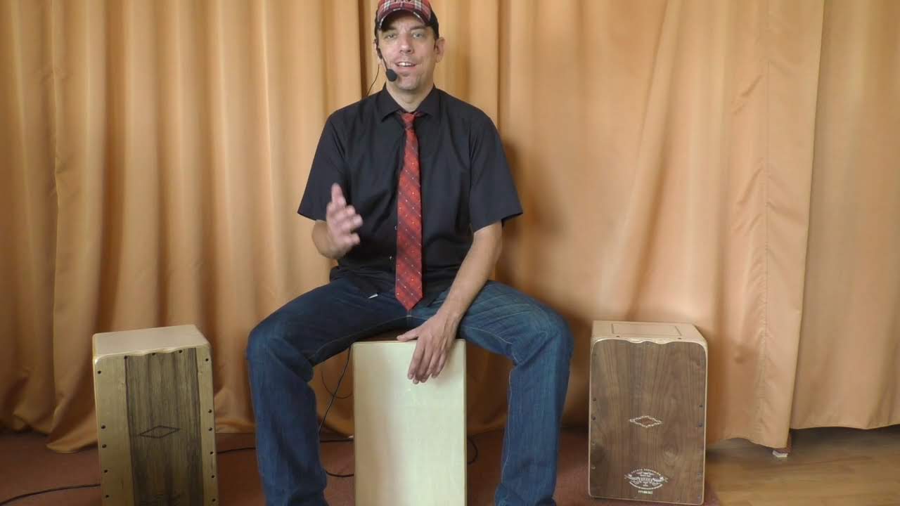 Cajon lernen – Percussion-Videokurs 3 Peter Lorson sitzt mit drei Cajons im Schulungsraum, trägt Mikrofon und erklärt etwas vor beiger Vorhangkulisse