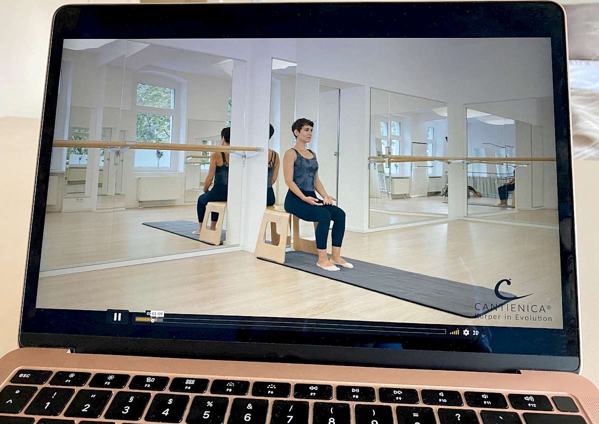 Videokurs-Screenshot: Frau sitzt auf Holzhocker vor Spiegeln mit Stange beim CANTIENICA® Training