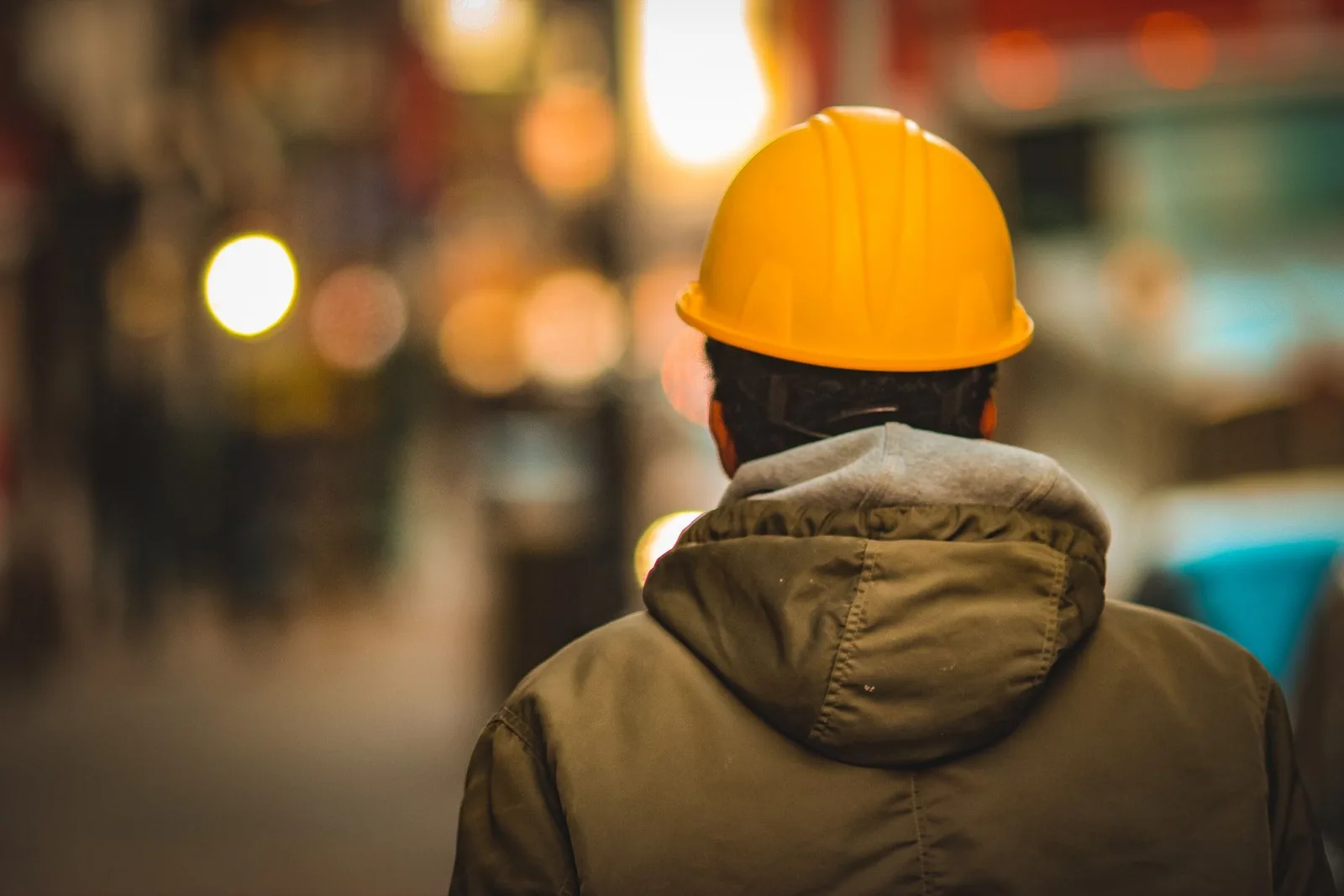 Person mit gelbem Schutzhelm und olivgrüner Jacke von hinten, Baustelle mit Lichtern