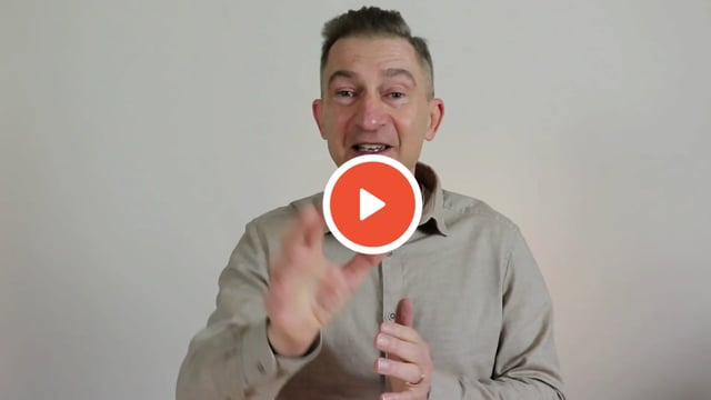 Videostandbild: Mann in beigem Hemd spricht gestikulierend vor weißem Hintergrund, Play-Symbol überlagert