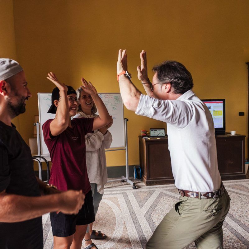 CEO Growth Mentoring – Coaching zur Unternehmensskalierung 7 Fünf Personen geben sich High-Fives in gelbem Seminarraum mit Whiteboards im Hintergrund