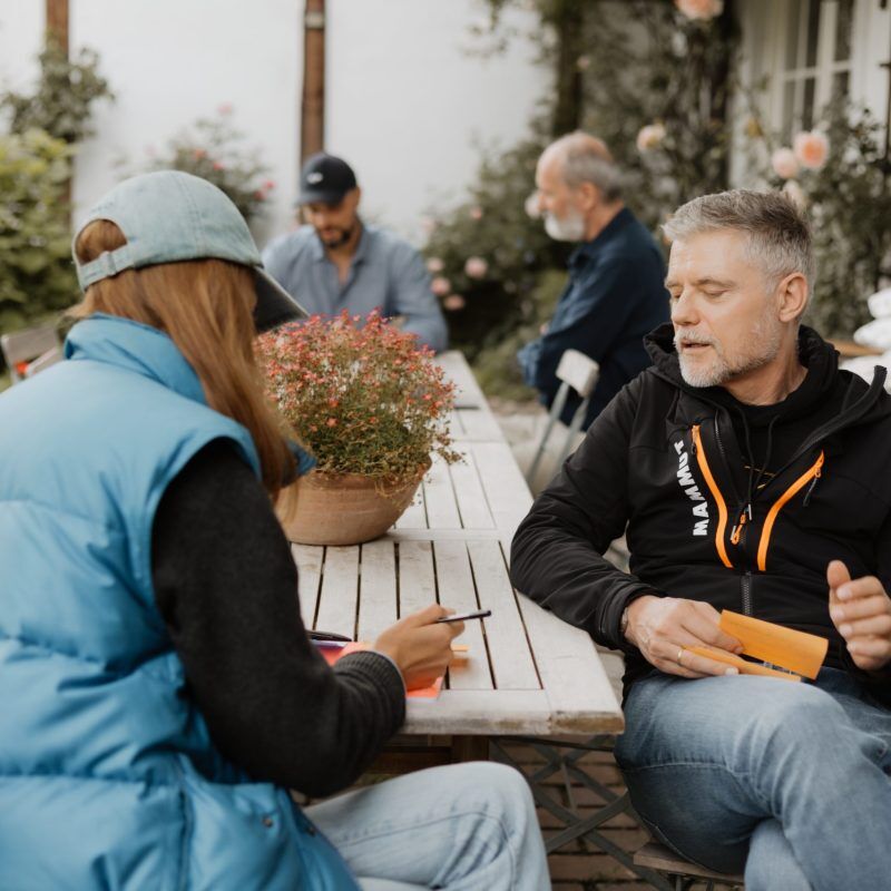 CEO Growth Mentoring – Coaching zur Unternehmensskalierung 10 Zwei Personen sitzen auf Terrasse beim Gespräch, Mann erklärt etwas, Frau hört zu und notiert