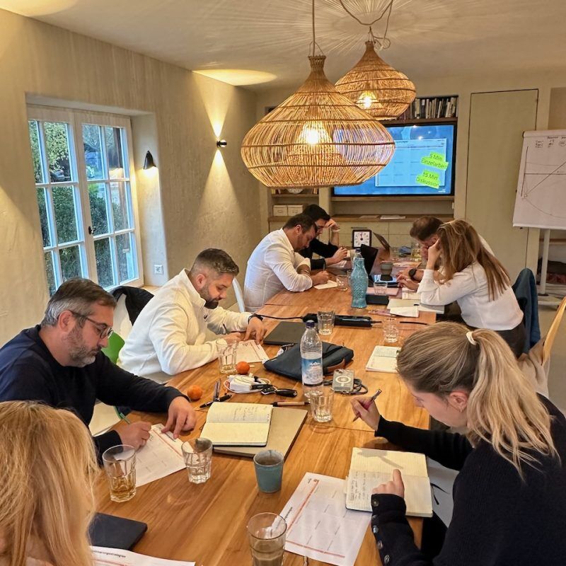CEO Growth Mentoring – Coaching zur Unternehmensskalierung 1 Mehrere Unternehmer sitzen an Holztisch in modernem Seminarraum mit hängenden Leuchten und Whiteboard