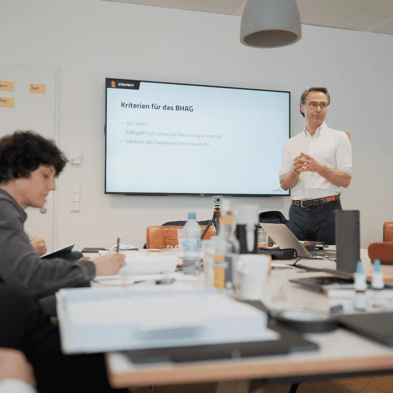 CEO Growth Mentoring – Coaching zur Unternehmensskalierung 20 Coach präsentiert Strategie vor Bildschirm mit Text 'Kriterien für das BHAG'