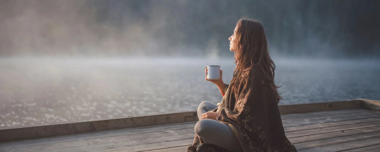 Frau sitzt auf Holzsteg am Wasser mit Kaffeetasse, neblige Seekulisse im Hintergrund