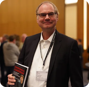 Charismatic Leadership – Praxisbuch über Führung 5 Mann Ralf J. mit Brille im dunklen Anzug hält das Charismatic Leadership-Buch
