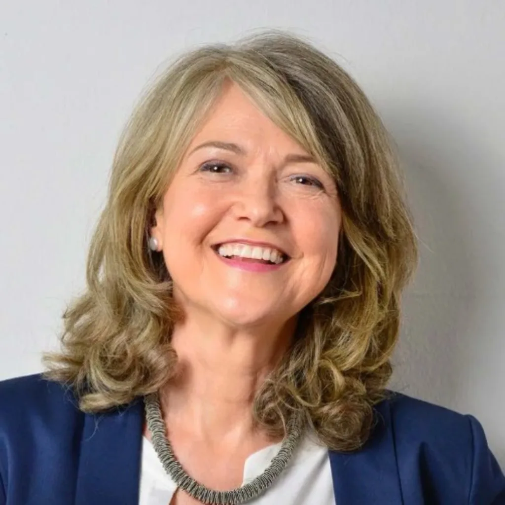 Porträt einer Frau mittleren Alters mit blondem Haar, dunklem Blazer und grauem Schal, lächelnd