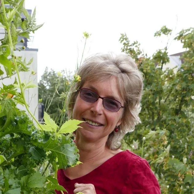 Chlorophyllkongress – Videokongress über Blattgrün von Maria Kageaki 4 Porträt einer Frau mit Brille und kurzen graumeliierten Haaren, lächelnd, vor Gartenkulisse mit Pflanzen