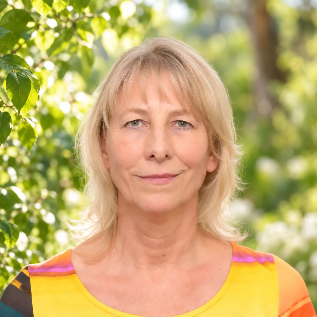 Chlorophyllkongress – Videokongress über Blattgrün von Maria Kageaki 29 Porträt einer Frau mittleren Alters mit blonden Haaren und orangegelbem Shirt vor grünem Blätterhintergrund