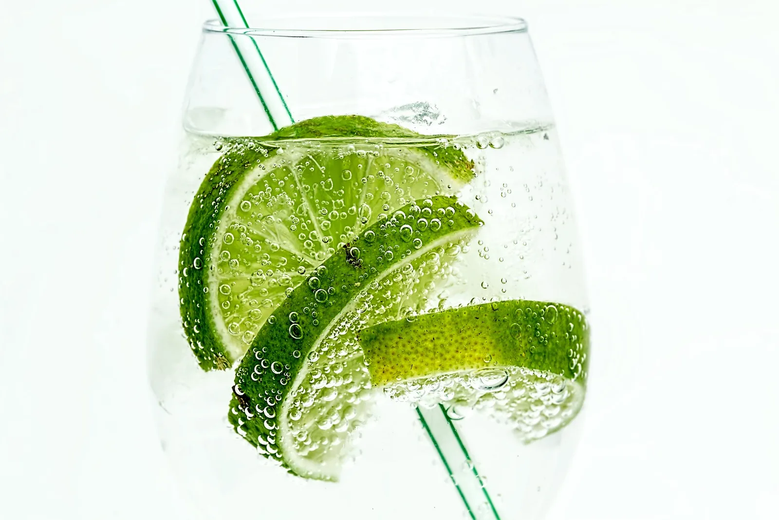 Glas mit Limettenscheiben und grünen Strohhalmen in Wasser mit Kohlensäurebläschen