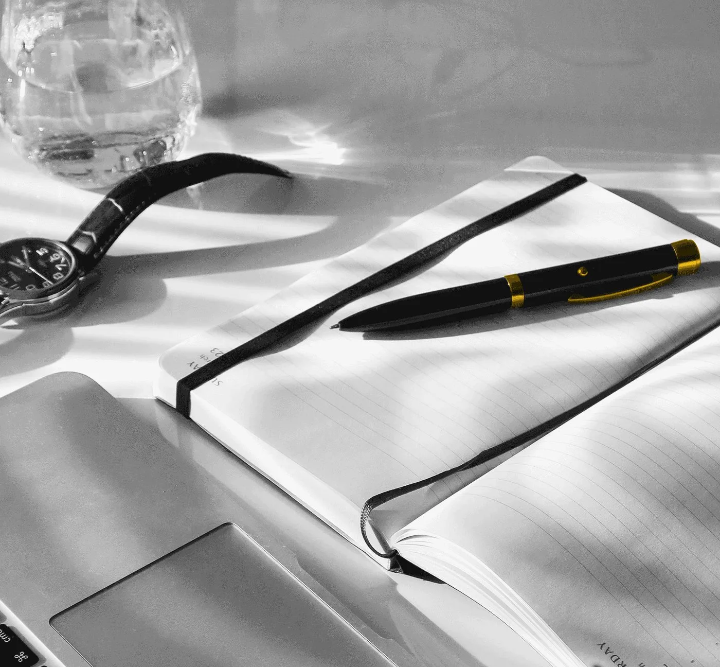 Schreibtisch mit Notizbuch, schwarzem Stift mit Goldakzenten, Uhr und Wasserglas