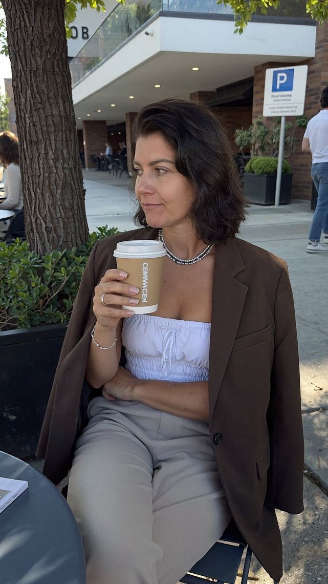 Frau in braunem Blazer und hellblauer Bluse hält Coffee-to-go Becher, steht vor modernem Gebäude