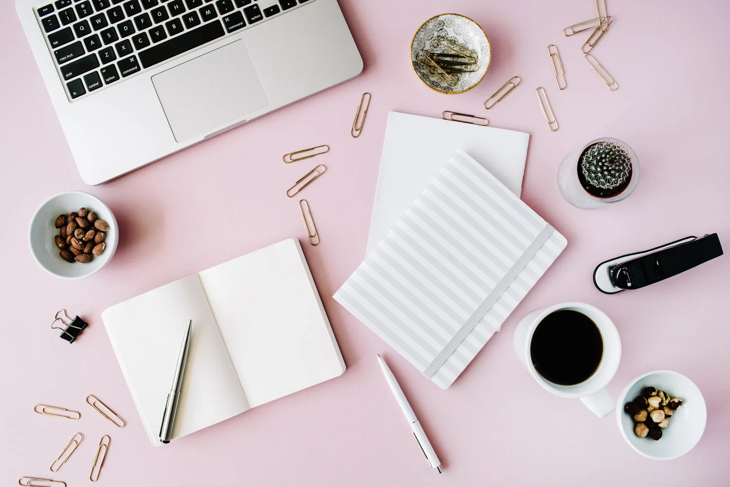 Schreibtisch-Flatlay mit Laptop, Notizbüchern, Stift, Kaffee und Pflanzen auf rosa Hintergrund