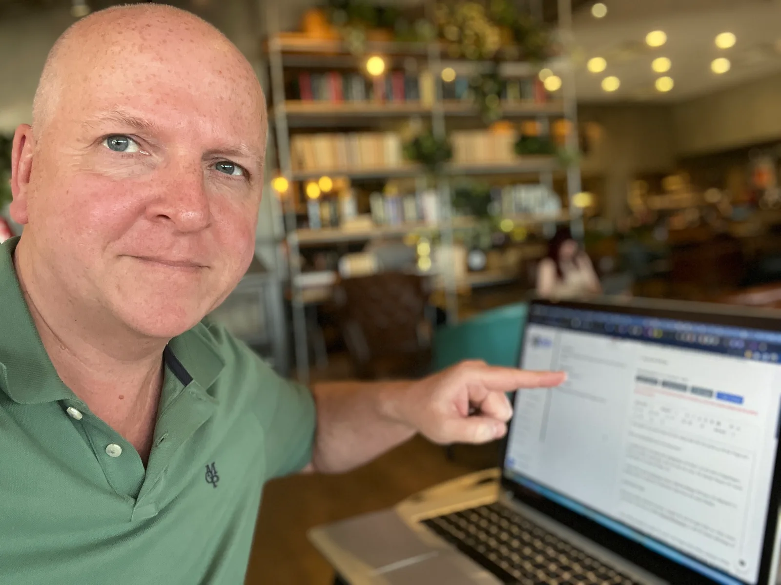 Mann in grünem Hemd am Laptop in modernem Büro mit Bücherregalen, pointing gesture