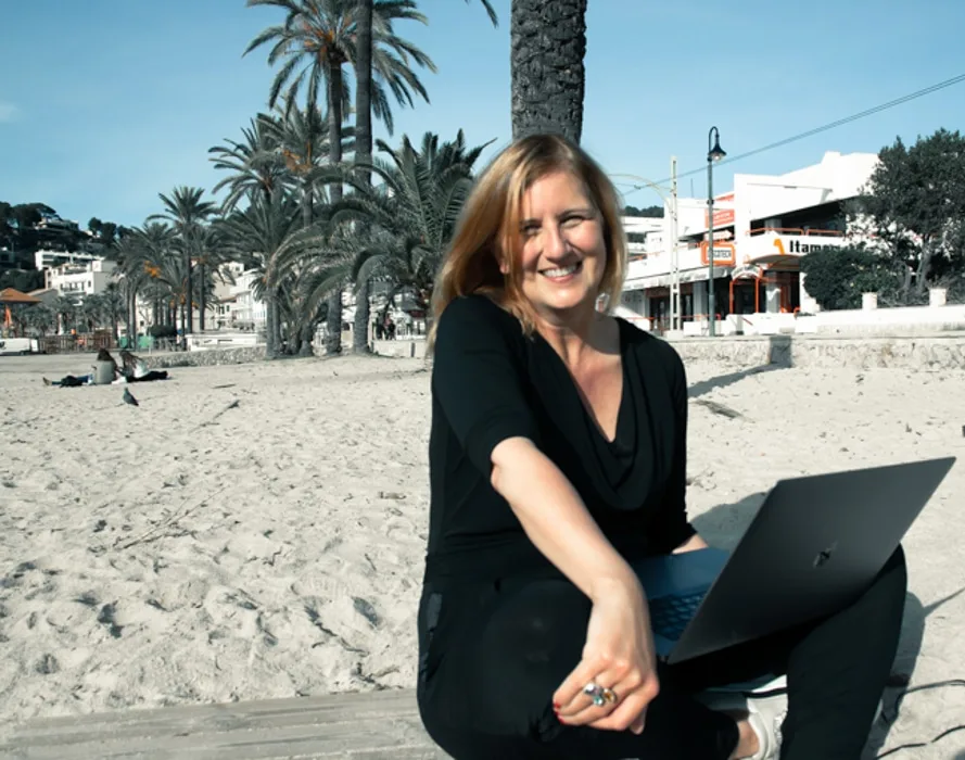 Beate Roth sitzt am Strand mit Laptop, schwarze Bluse, braune Haare, lächelnd
