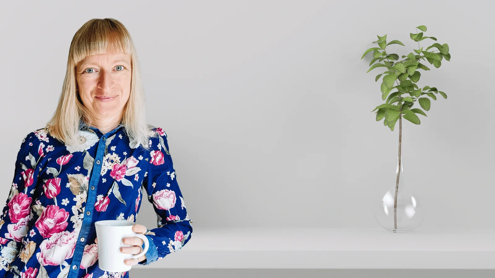 Frau mit blondem Haar und Brille in blauer Blüten-Bluse hält Kaffeetasse, neben Pflanze in Vase