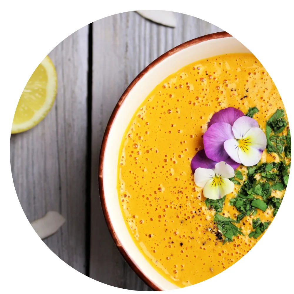 Gelbe Creme-Suppe in Schale mit Passionsfrüchten, Blüten und Kräutern, Zitronenscheibe