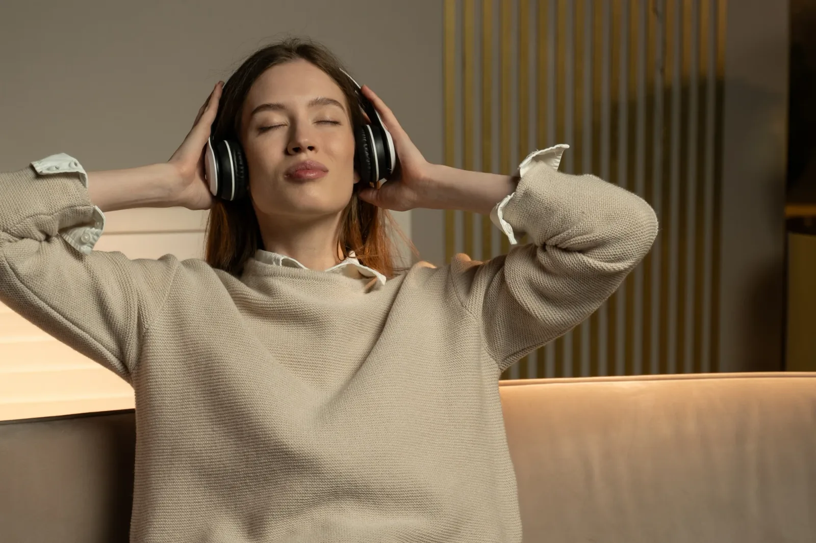 Frau mit Kopfhörern lehnt sich zurück, Augen geschlossen, cremefarbenes Sweatshirt, warmes Studioambiente