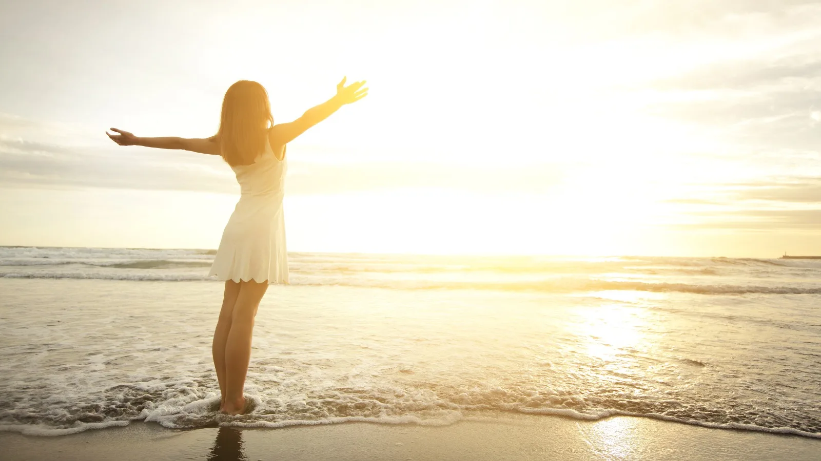 Frau in weißem Kleid mit ausgebreiteten Armen am sonnigen Strand bei Sonnenaufgang