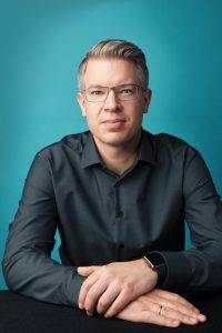 Portrait eines Mannes mit kurzen Haaren und Brille vor blauem Hintergrund
