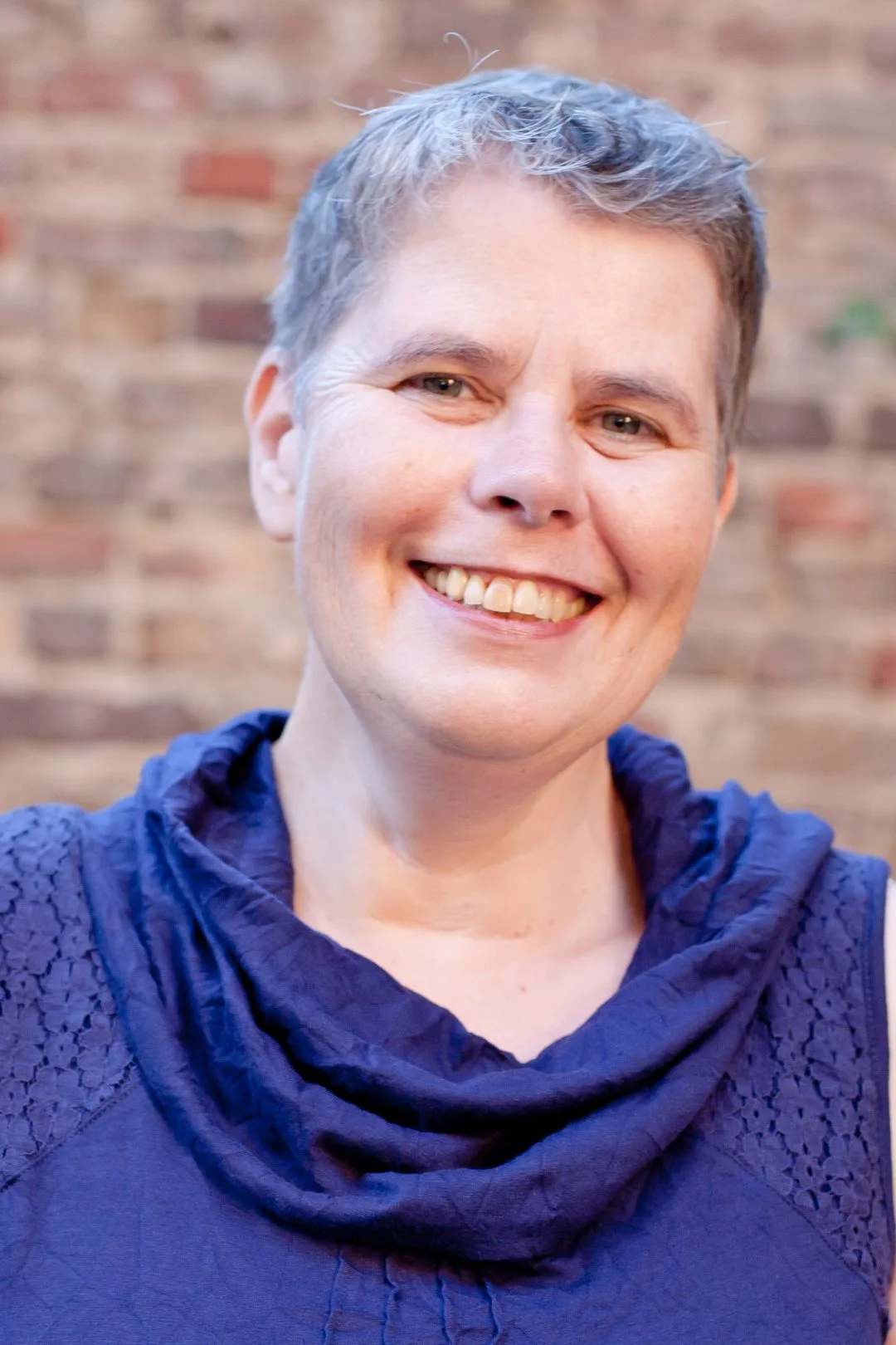 Protraitfoto von Monika Richrath lächelnd vor Ziegelmauer mit blauem Oberteil