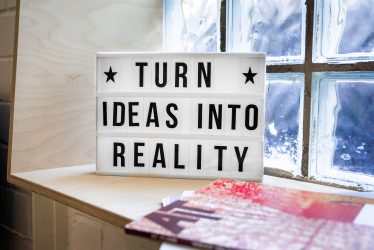 Lightbox mit Text Turn Ideas Into Reality auf Schreibtisch mit rosa Notizbuch und Fenster