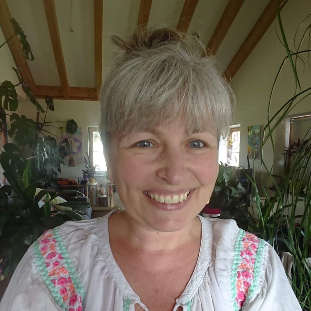 Porträt einer Frau mit kurzen grauen Haaren und Pony, lächelnd in Innenraum mit Pflanzen, Bluse mit Blumenmuster