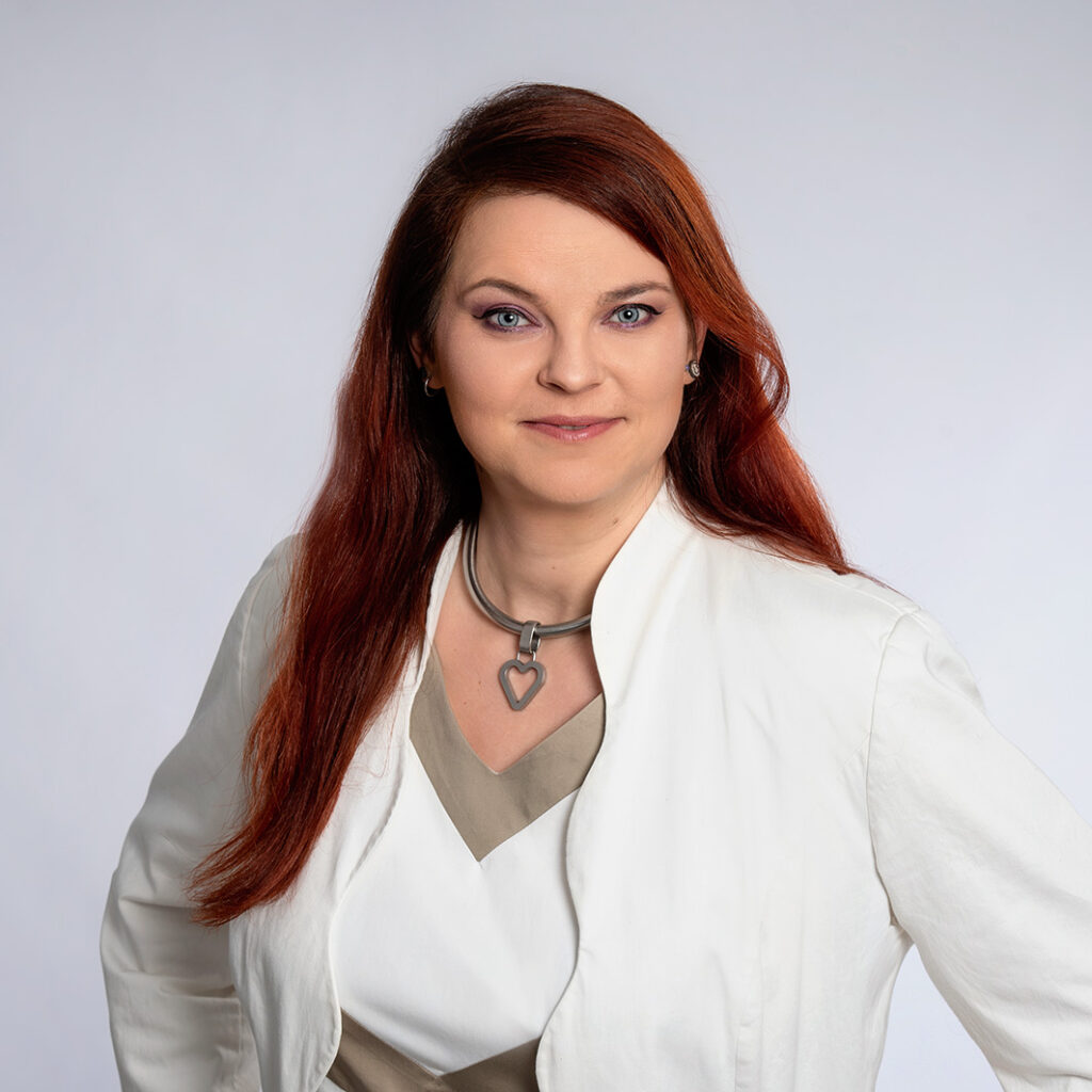 Frau mit langen rötlichen Haaren, weißer Blazer, Herz-Halskette, neutraler grauer Hintergrund