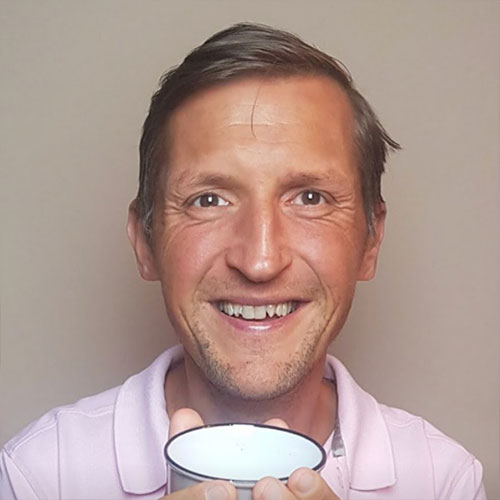 Mann mit rosa Hemd, kurzen dunklen Haaren, lächelt in die Kamera, neutraler Hintergrund