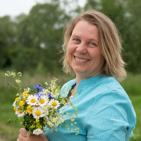 Heike Engel lächelt in die Kamera, trägt hellblaues Hemd, hält Strauß mit Gänseblümchen und blauen Blüten