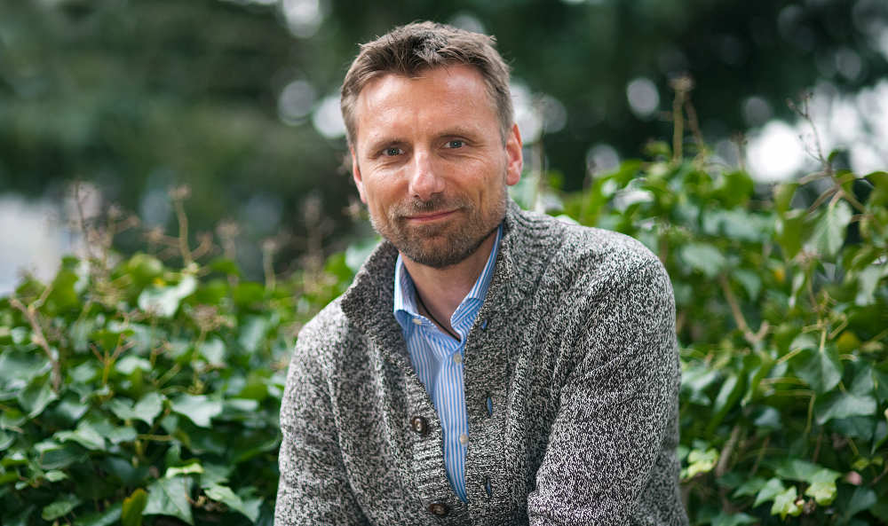 Porträt von René Penselin lächelnd in grauem Pullover vor Garten-Hintergrund