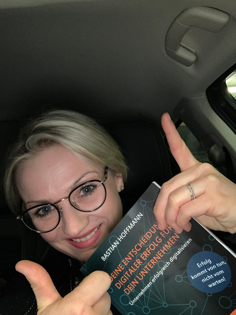 Junge Frau mit Brille im Auto hält Buch nach vorne, lächelt