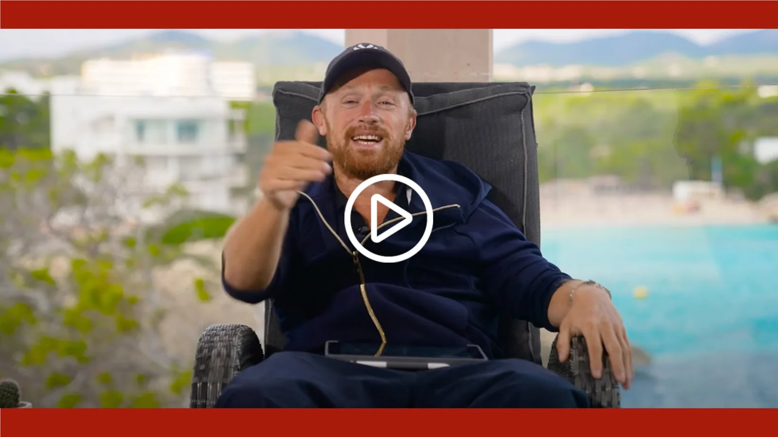 Videostandbild von Danny Adams mit schwarzer Kappe lachend in Stuhl sitzend vor Landschaft mit Play-Symbol