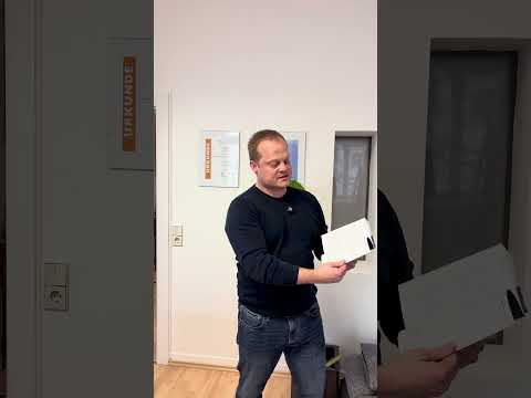 Bernd Krause steht in hellem Flur mit Buch in der Hand, trägt dunklen Pullover, lächelt in Kamera
