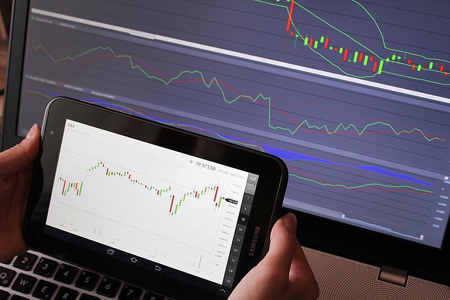 Hände halten Smartphone mit Candlestick-Charts, Monitor im Hintergrund mit Handelsgrafen