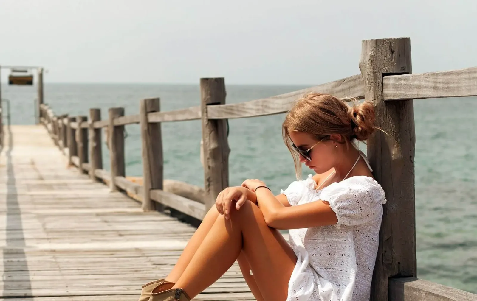 Junge Frau sitzt auf Holzsteg am Meer, trägt Sonnenbrille und weißes Kleid, schaut aufs Wasser