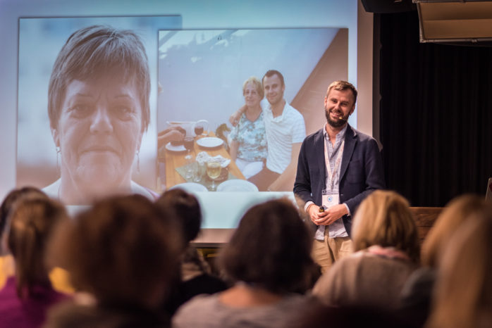 Mann präsentiert auf Bühne vor Publikum mit Beamerpräsentation im Hintergrund