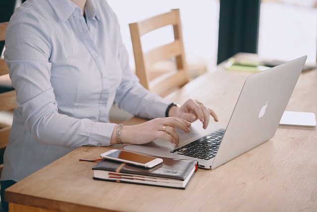 Frau in weißer Bluse tippt auf MacBook am Schreibtisch mit Notizbuch und Smartphone