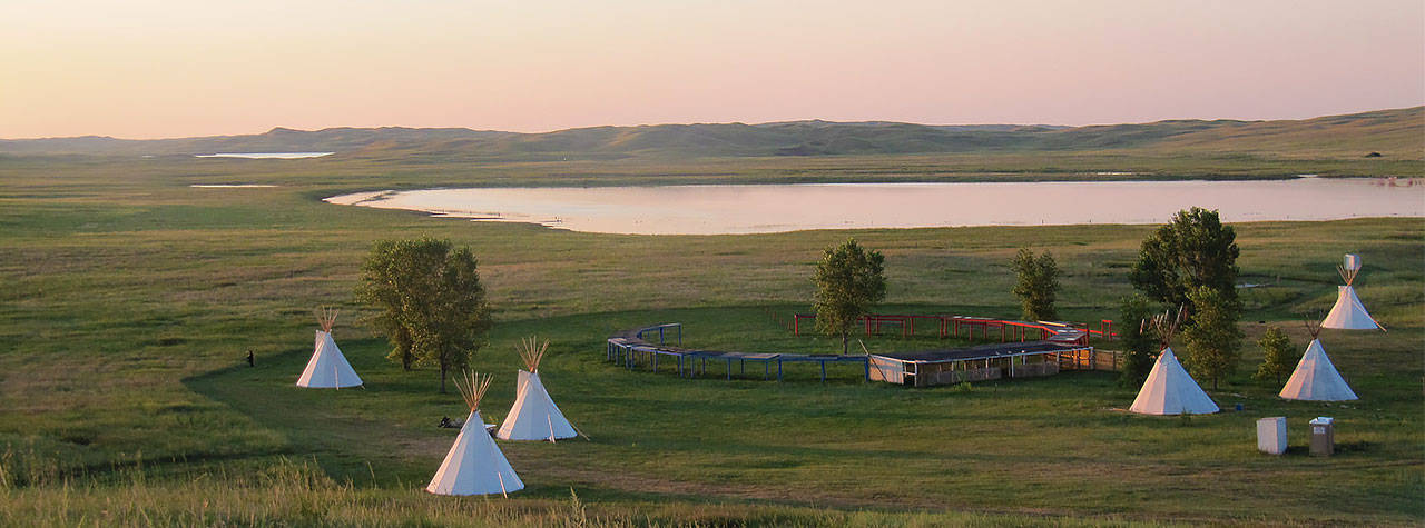 Mehrere weiße Tipis auf grüner Wiese mit See und Bergen im Hintergrund bei Sonnenuntergang