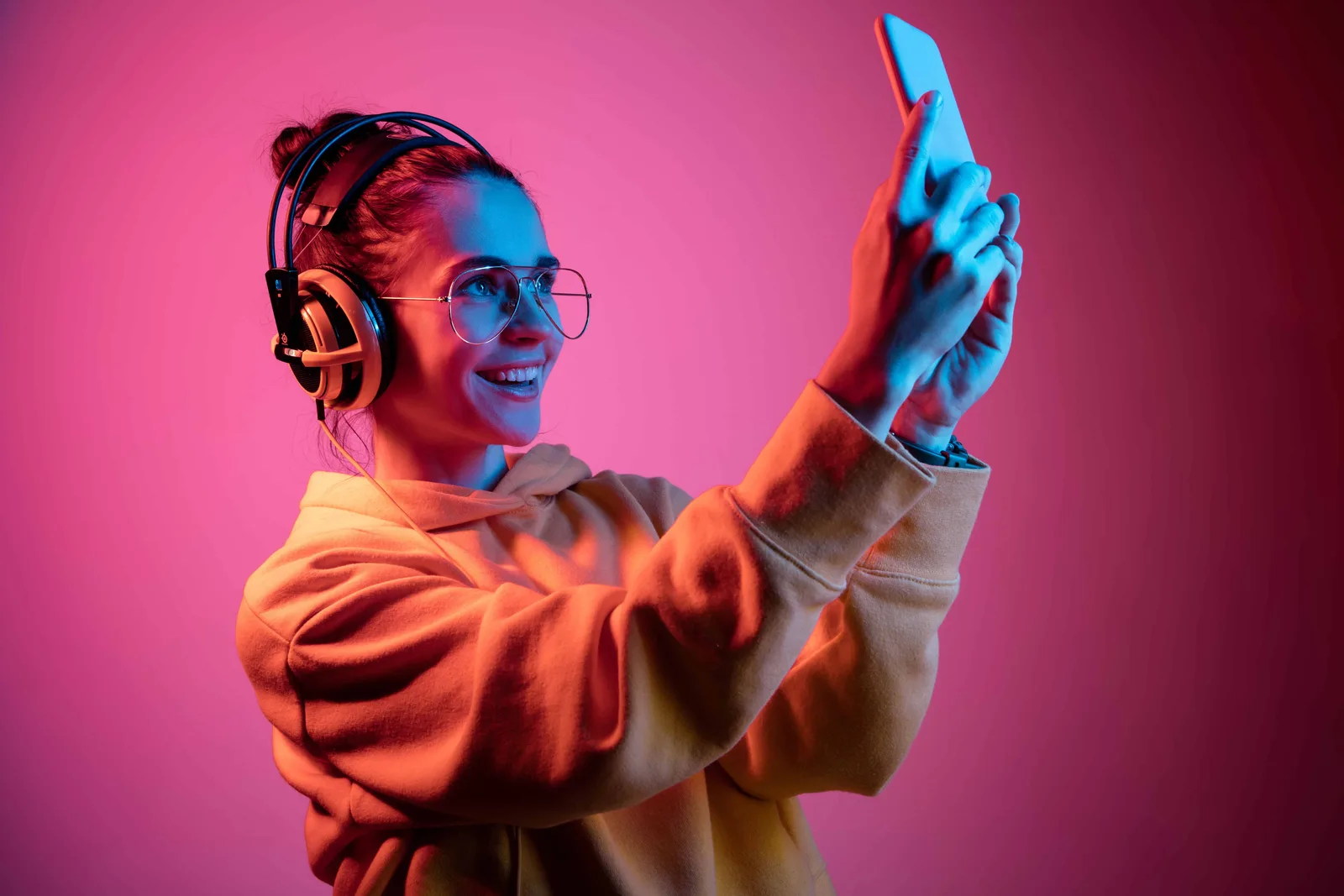 Junge Frau mit Kopfhörern, Brille und Smartphone vor pink-blauem Hintergrund