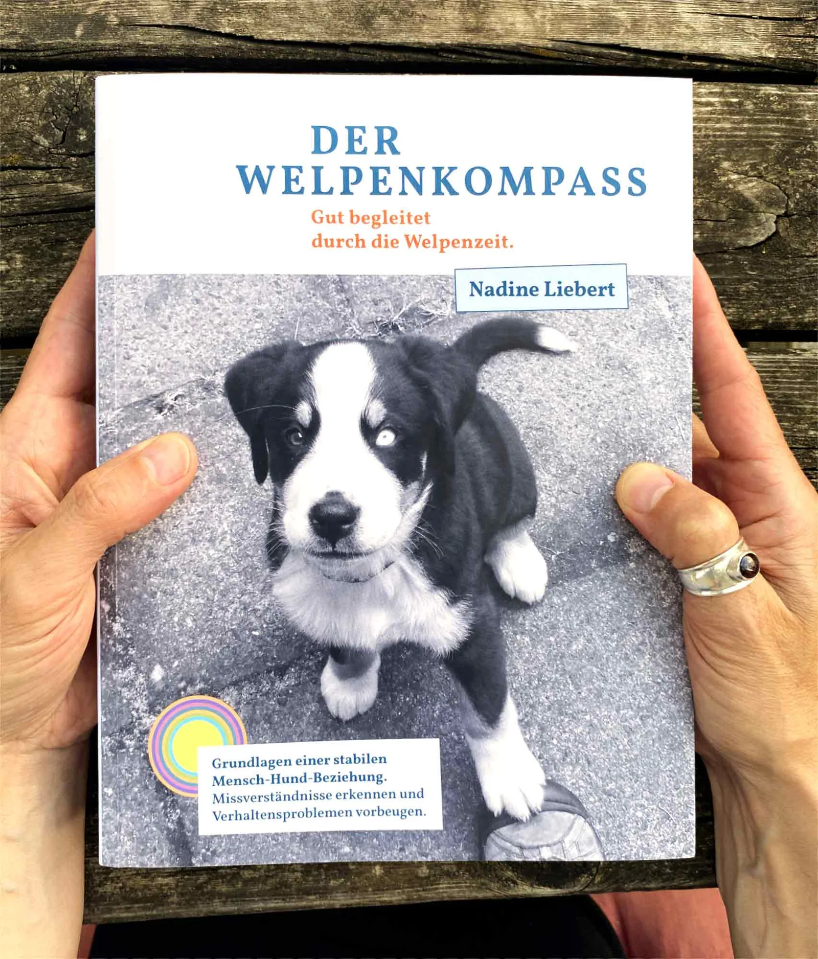 Der Welpenkompass – Praxisbuch zur Welpenzeit von Nadine Liebert 5 Buchcover Der Welpenkompass in Händen vor rustikalem Holzhintergrund
