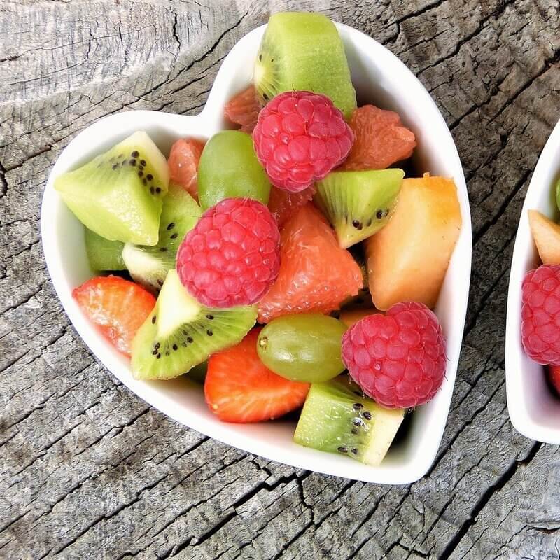 Herzförmiger weißer Teller mit frischen Früchten: Kiwi, Erdbeere, Himbeere, Traubenkernöl auf Holzuntergrund