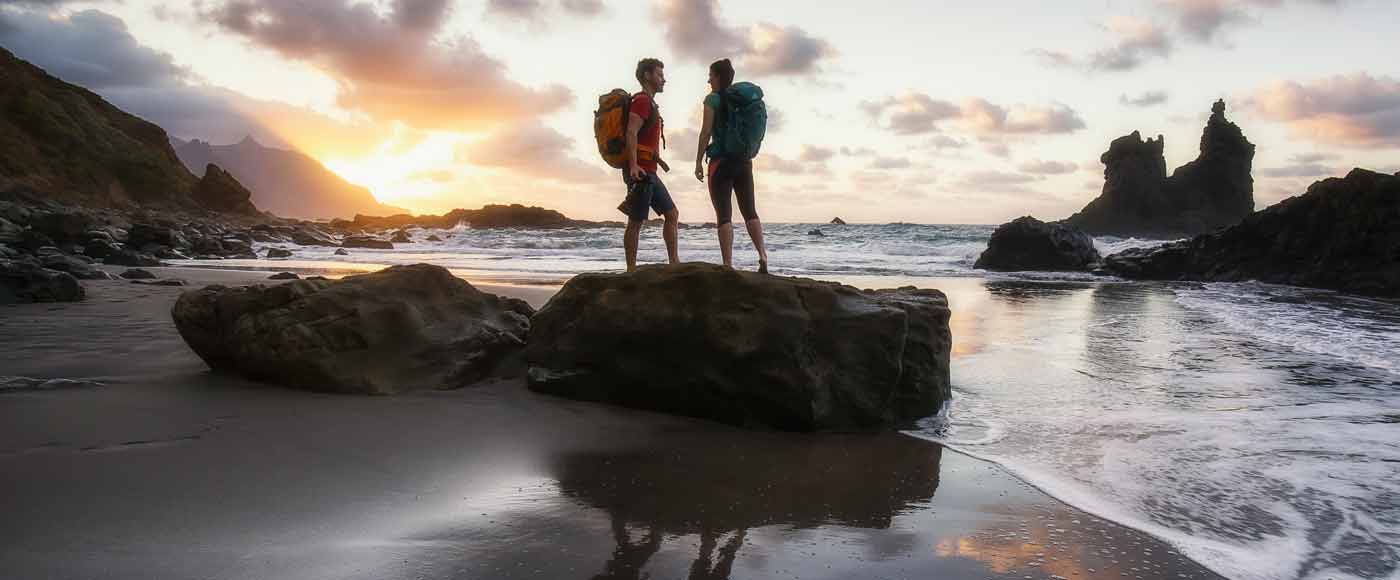 Zwei Wanderer mit Rucksäcken auf Felsen am Strand bei Sonnenuntergang, Wellen und Klippen im Hintergrund
